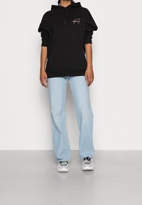 Sudadera negra con capucha, bolsillo frontal y logo en el pecho, combinada con jeans de campana de color azul claro y zapatillas grises sobre un suelo gris.