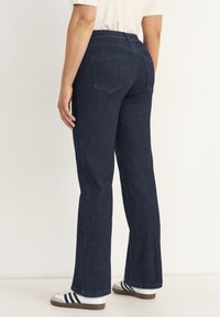 Kvinna i mörkblå raka jeans och vita sneakers med svarta ränder mot en enfärgad bakgrund.