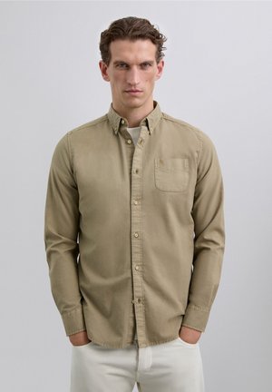 Hombre con cabello rizado corto, vistiendo una camisa beige de botones sobre una camiseta blanca, con las manos en los bolsillos, de pie contra un fondo gris claro liso.