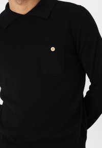 Maglione nero a maglia con colletto polo, dotato di una tasca sul petto decorata con un bottone di colore chiaro. Texture morbida, vestibilità aderente.