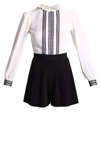 Blusa blanca con cuello rizado y detalle de encaje negro, combinada con pantalones cortos negros de cintura alta con pliegues. Mangas largas con puños acentuados.