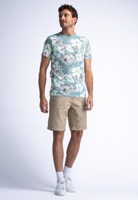 Lichtblauw bloemen T-shirt met witte en groene accenten, gecombineerd met kaki shorts. Schoeisel bestaat uit witte sneakers met witte sokken.