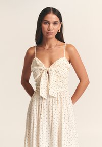 Cream dress with black polka dots, featuring a tied front, adjustable spaghetti straps, and a flowing skirt. Smooth, lightweight fabric.