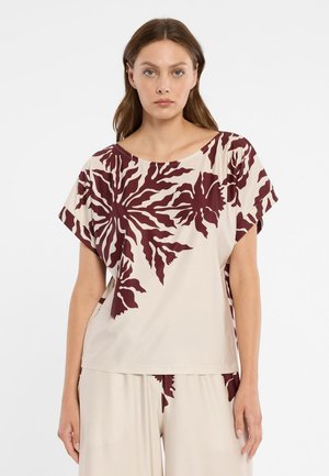 Vrouw draagt een beige top en broek met grote donkerrode abstracte bladpatronen, staand tegen een effen witte achtergrond.