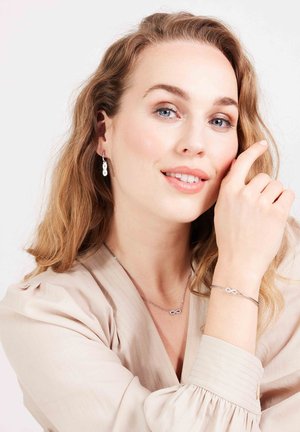 Zilveren sieraden met een oneindigheidssymbool op een delicate armband en bijpassende ketting, gedragen met een blouse in neutrale tinten.