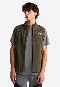 Salmivihreä pehmeä kuoriliivi pystykauluksella, edessä vetoketju ja logotäyte. Päällä harmaa poolopaita, sivutaskut.