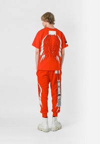 Camiseta roja con gráficos de alas blancas en la parte posterior, combinada con pantalones rojos a juego que tienen impresiones de alas en los laterales. Llevada con botas blancas hasta el tobillo.