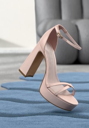 Világos rózsaszín magassarkú szandál bokapánttal és platformtal, kék szőnyegen elhelyezve, háttérben elmosódott szürke kanapéval.