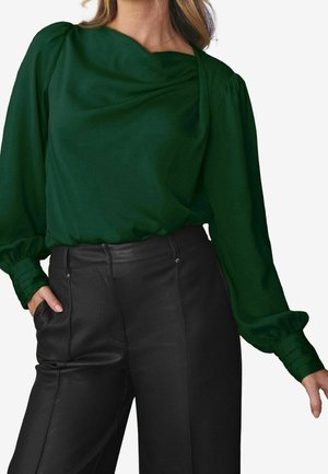 Frau trägt eine dunkelgrüne Bluse mit langen Ärmeln und plissierten Bündchen sowie schwarze, hoch taillierte Lederhose, eine Hand in der Tasche.