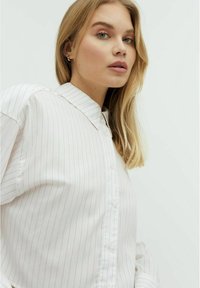 Camicia bianca con delicate strisce verticali rosa, dotata di colletto classico e vestibilità rilassata. Tessuto di cotone liscio e maniche arricciate.