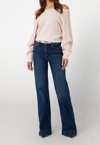 Helles rosa Off-Shoulder-Pullover mit strukturiertem Strick, kombiniert mit hochgeschnittenen dunkelblauen Schlagjeans und einem rosa Gürtel. Schlichte Perlenkette.