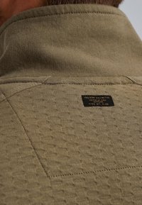 Olijfgroen pullover met een gestructureerd patroon, voorzien van een hoge kraag en een zwart label met reliëftekst dat op de achterkant is genaaid.