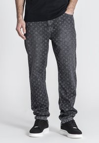 Jeans de mezclilla negra con un patrón de monograma en tono sobre tono, corte recto y costuras en contraste. Llevados con zapatillas negras.