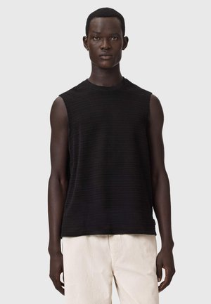 Homme à la peau foncée portant un haut noir sans manches texturé et un pantalon beige clair, debout devant un fond uni gris clair.