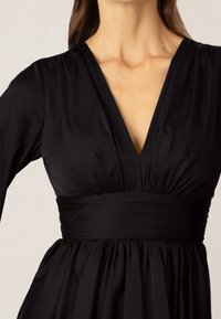 Svart klänning med djup V-ringning, långa ärmar och gathered midja. Slät textur med figurnära överdel och flödande kjol.