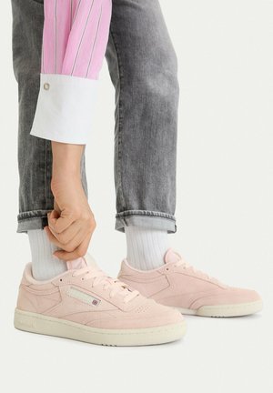Person in grauen Jeans und weißen Socken zieht hellrosa Reebok-Sneaker mit einer weißen Sohle vor einem schlichten Hintergrund an.