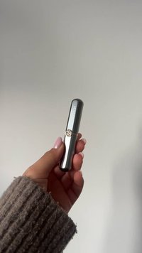 Czarna szminka w tubce z błyszczącym wykończeniem, złotym akcentem i okrągłym emblematem. Trzymana w ręce ubrane w prążkowany, teksturowany sweter.