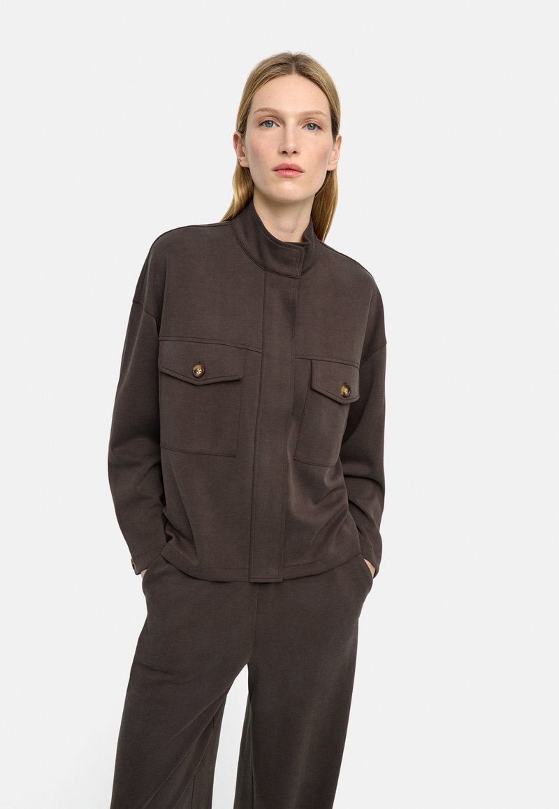 Kobieta o jasnych włosach ubrana w ciemnobrązową kurtkę z długimi rękawami i zapinanymi na guziki kieszeniami na piersi oraz pasujące szerokie spodnie, ręce w kieszeniach.