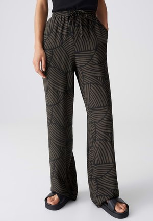 Personne portant un pantalon large noir et marron à motif abstrait avec taille à cordon et sandales plateformes noires, mains dans les poches.