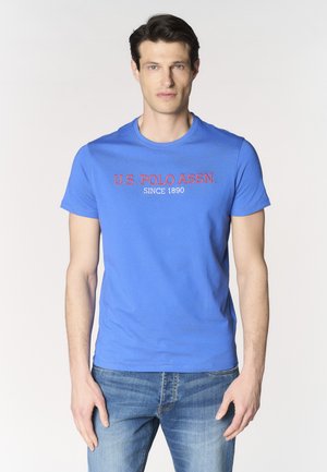 Uomo che indossa una t-shirt blu U.S. Polo Assn. e jeans blu, in piedi davanti a uno sfondo chiaro e semplice.