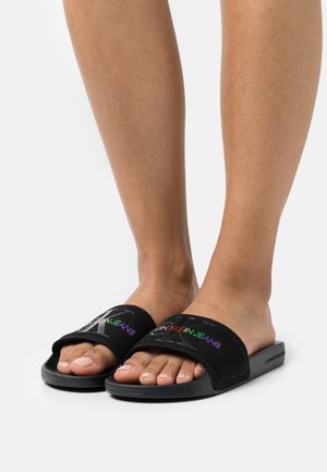 Czarne sandały na korek z materiałowym paskiem z napisem „CK” w wielokolorowych literach. Teksturowana podeszwa i otwarty nosek dla wygody.