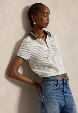 Γυναίκα με κοτσίδες, φορούν λευκό πουλόβερ πόλο με πολύχρωμη τρέσα και μπλε τζιν, στέκεται μπροστά σε λευκό τοίχο με κλειστά μάτια.