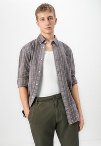 Jovem com cabelo curto, vestindo uma camisa de xadrez com mangas arregaçadas sobre uma camisola branca e calças verde-oliva, contra um fundo branco.