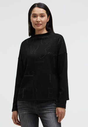 Frau mit glattem schwarzen Haar, die ein schwarzes Langarmshirt mit dezenten Buchstabenmustern und ausgewaschene graue Jeans trägt, steht vor einem schlichten Hintergrund.