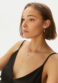Collana d'argento con un pendente curvo, indossata sopra un top senza maniche nero; la modella ha i capelli corti e indossa un orecchino di perle.