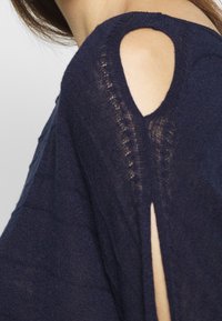 Marinblå tröja med urklippta axeldetaljer, strukturerad stickad tyg och en avslappnad passform. Sidofickor förbättrar andningsförmågan.