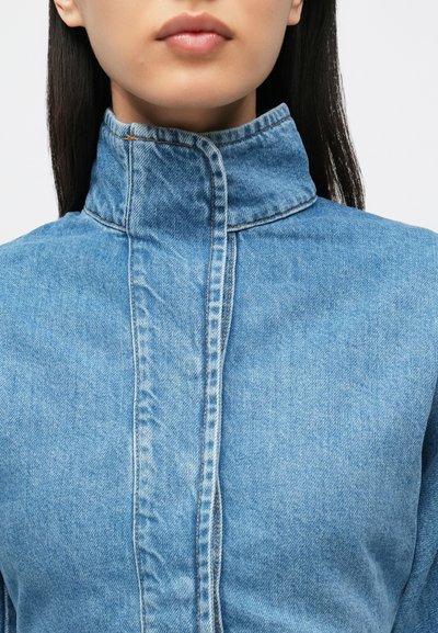 Gaiši zila džinsa jaka ar augstu apkakli, centrālu šuves detaļu un kontrastējošu šuvju rakstu. Audums ir gluds tekstūrā.