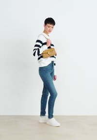 Striped black-and-white sweater, blue skinny jeans, white sneakers, holding a quilted, tan handbag with a chain strap, minimal background.