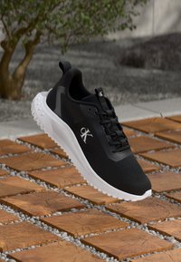 Schwarzer Calvin Klein Sneaker mit weißer Sohle, schwebend über einem Boden aus Holzfliesen und Kies, im Freien in der Nähe eines Baumes.