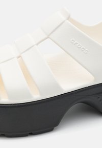 Fehér Crocs szandál fekete platformtalppal, több vízszintes kivágással a pánton. Texturált felsőrésze és sima talpa van.