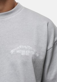 Sweat-shirt gris clair avec un col ras du cou. Doté d'un texte brodé blanc indiquant "GOOD FOR NOTHING BRITISH ATELIER EST 2011." Texture douce.