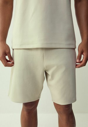 Man wearing light beige casual shorts and matching shirt, standing with hands relaxed by sides.