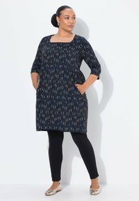 Vrouw met handen in de zakken, gekleed in een zwarte gebloemde jurk tot de knie, zwarte leggings, metallic ballerina's en gouden oorringen, staande tegen een witte muur.
