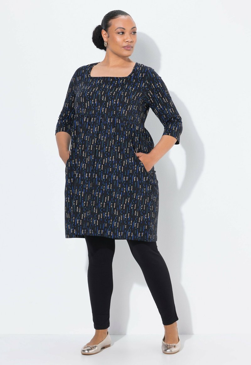 Donna con le mani in tasca, che indossa un abito nero con motivo fino al ginocchio, leggings neri, ballerine metalliche e orecchini a cerchio dorati, in piedi contro un muro bianco.