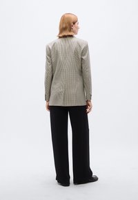 Καρό σακάκι σε μπεζ και μαύρο, με δομημένη εφαρμογή, μακριά μανίκια και σκίσιμο στο πίσω μέρος, συνδυασμένο με φαρδιά μαύρη παντελόνα.