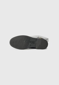 Semelle en caoutchouc noir d'une botte avec un motif texturé, dotée d'un logo en relief et d'une partie supérieure métallique lisse avec une fermeture éclair.