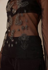 Black sheer top featuring intricate silver embroidery of butterflies, flowers, and celestial motifs. Finished with frayed edges and a button closure.