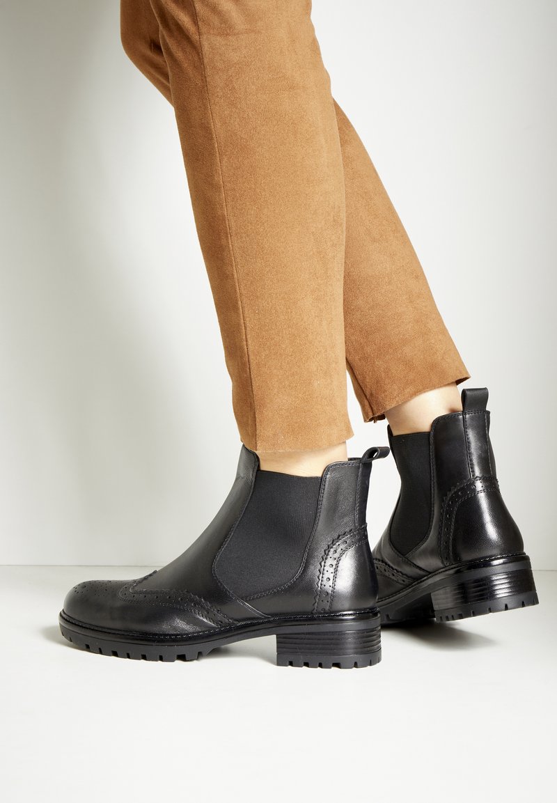 Zwarte leren enkelboots met elastische zijpanelen, brogue-details en een stevige gestructureerde zool, gecombineerd met tan suède broek.