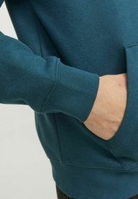 Sudadera de color teal con puños y dobladillo acanalados. Incluye un bolsillo canguro y una textura de tela suave, mostrando una mano descansando en el bolsillo.