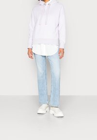 Sudadera con capucha color lila sobre una camisa blanca abotonada, combinada con vaqueros azul claro de pierna recta y botas blancas con suela gruesa.