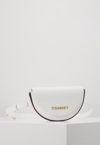 Bílá pásek taška ve tvaru půlměsíce z kůže se zlatým nápisem "TOMMY" a nastavitelným popruhem na bílém povrchu na jednobarevném pozadí.