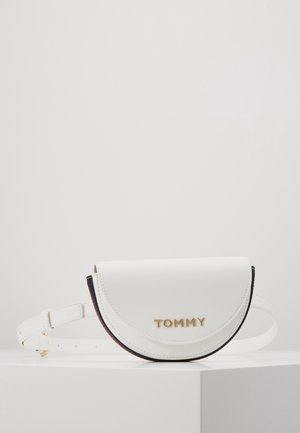 Sac à ceinture en cuir en forme de demi-lune blanche avec lettrage doré "TOMMY" et bandoulière réglable sur une surface blanche, contre un fond uni.
