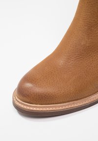 Hnědé kožené kotníkové boty s texturovaným povrchem, mají kulatou špičku a prošívanou hnědou podrážku. Boty mají hladký ochranný kryt špičky pro zvýšení trvanlivosti.
