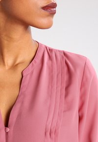 Close-up of a person wearing a dusty pink blouse with pleats near the neckline and glossy mauve lipstick.
