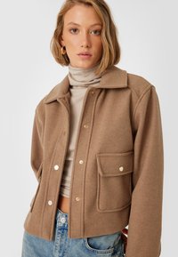 Giacca di lana marrone con colletto, bottoni a pressione dorati e tasca frontale. Indossata sopra un dolcevita beige e jeans a vita alta blu.
