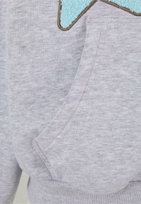 Pelēkas sporta krekliņa audums ar mīkstu tekstūru, kuram ir tirkīza krāsas flīsa zvaigznītes aplikācija un sānu kabata. Papildu detaļas ietver ribotās apakšmalu un aproces.